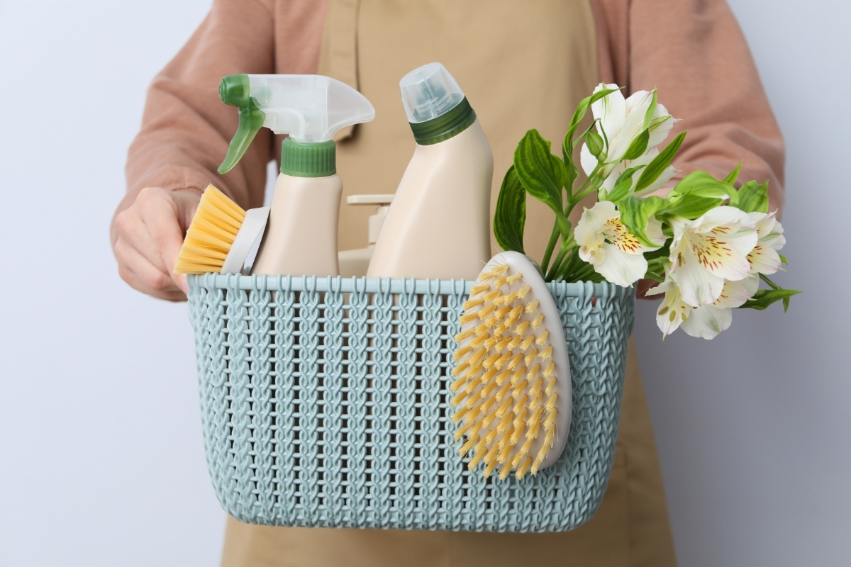 Blog Persoană care deține un coș albastru umplut cu sticle de spray de curățare, două perii de frecat, un burete, și un mic buchet de flori albe - perfect pentru cei care caută curatenie Bucuresti sau expert servicii curatenie.